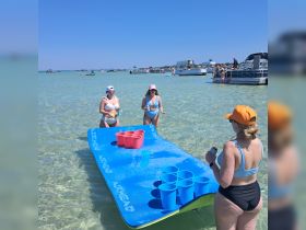 A lively group enjoys a game of beer pong on a floating mat in shallow waters, perfect for summer beach parties and fun under the sun.