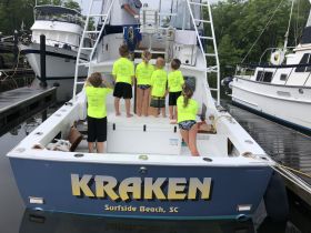 A group of children in bright yellow shirts eagerly explore the back of the 'Kraken' boat at the dock, showcasing the excitement of a day out on the water.