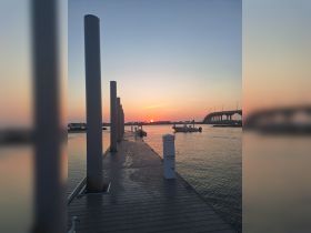 A picturesque sunset view from a dock, with a bridge in the background and a boat gently anchored nearby, creating a serene atmosphere perfect for evening strolls.