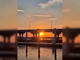 A serene sunset view reflecting off calm waters, framed by a bridge, creating a picturesque scene perfect for evening strolls or boat rides.