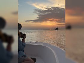 A person captures the stunning sunset while aboard a boat, with a fishing vessel in the distance, creating a picturesque scene perfect for photography enthusiasts.