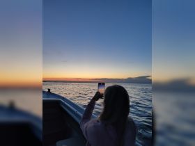 A woman captures the stunning sunset on her phone while enjoying a peaceful moment on the water, showcasing the beauty of a boat trip at dusk.