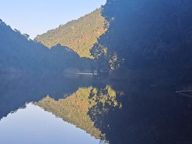 A serene landscape reflecting mountains and trees in calm waters, creating a tranquil atmosphere. The image quality is unclear, making it difficult to discern details.