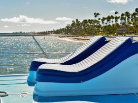 A cozy seating area on a boat, featuring comfortable lounge chairs with a stunning view of the shimmering water and palm trees in the background — perfect for relaxing on a sunny day.