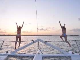 Two friends joyfully jump off the front of a catamaran into the water at sunset, capturing the essence of fun and adventure on a perfect boating trip.