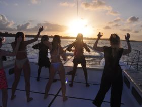 A lively group dances on the deck of a catamaran at sunset, celebrating with drinks and enjoying the beautiful ocean views — perfect for unforgettable party experiences on the water.