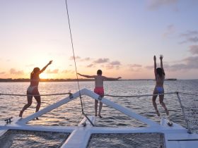 A joyful group of friends jumps off the front of a catamaran at sunset, capturing the essence of fun and adventure on the water.
