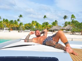 A man relaxes on the deck of a boat, soaking up the sun with a beautiful beach backdrop — perfect for a day of leisure and enjoyment on the water.