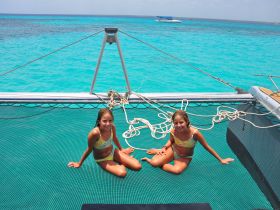 Two girls enjoy a sunny day on a catamaran, relaxing on the netting above the stunning turquoise waters — a perfect moment for fun and adventure on the water.