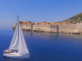 A stunning aerial view of a sailboat gliding through calm waters near a historic coastal town, showcasing the beautiful architecture and vibrant rooftops against a clear blue sky.