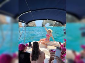 A lively group enjoys a fun day on the water, with one guest posing in a pink bikini while others capture the moment on their phones. The stunning coastal scenery adds to the festive atmosphere.