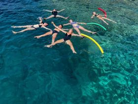 A lively group enjoys a fun day swimming in crystal-clear waters, using colorful pool noodles for added enjoyment — perfect for summer adventures on the water.