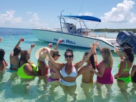 A lively group of friends celebrates in the shallow waters, raising cocktails with a boat in the background — perfect for a fun day on the water.