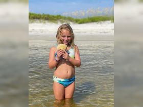 A young girl joyfully holds a shell while standing in shallow water, capturing a delightful moment of beach exploration and wildlife interaction.