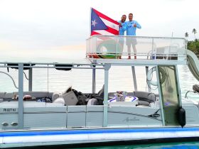 Two friends celebrate aboard a pontoon boat, proudly displaying a flag against a serene water backdrop — perfect for group outings and fun on the water.