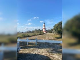 A scenic view of a lighthouse with people walking along a path, set against a clear blue sky. The image captures a tranquil outdoor setting, ideal for exploring coastal landmarks.