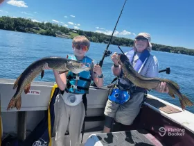 A young boy and an older man proudly display their catches of pike while fishing on a sunny day, showcasing the joy of outdoor fishing adventures.