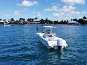 A person stands on a fishing boat in clear blue waters, showcasing an active day on the water amidst a scenic backdrop of palm trees and waterfront homes.