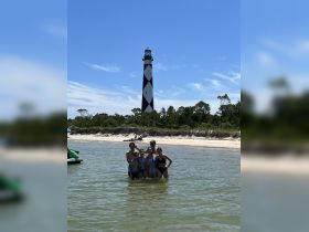 A cheerful group enjoys a sunny day at the beach, posing in shallow water with a striking lighthouse in the background — perfect for family outings and summer adventures.