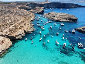 A stunning aerial view of numerous boats anchored in crystal-clear turquoise waters, surrounded by rocky cliffs and beachgoers enjoying the sun — a perfect destination for island hopping adventures.