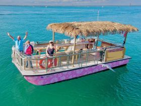 A cheerful group waves from a colorful tiki boat, enjoying a sunny day on the water — perfect for fun outings and celebrations.
