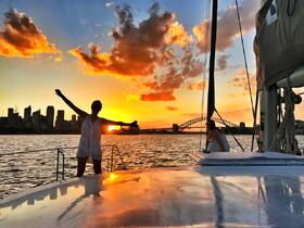 Two women enjoy a stunning sunset aboard a sailboat, with the city skyline and iconic bridge in the background — a perfect moment for sailing enthusiasts and sunset lovers.