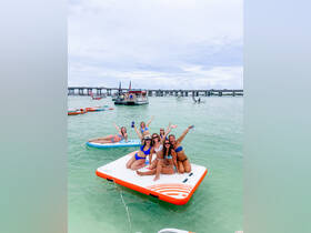A lively group of friends enjoys a fun day on the water, celebrating on a floating mat while surrounded by beautiful scenery and a pontoon boat in the background.