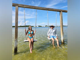 A couple enjoys a playful moment on swings in shallow water, surrounded by a beautiful beach backdrop — perfect for summer fun and memorable experiences.