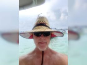 A blurry selfie of a person wearing a straw hat and sunglasses, with a water background. The image lacks clarity, making it difficult to discern details.