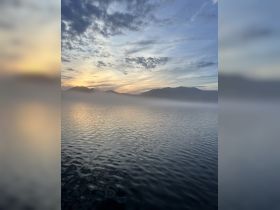 A serene view of a misty lake at sunset, with soft clouds and mountains in the background, creating a tranquil atmosphere.