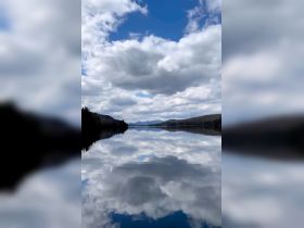 A serene view of a tranquil lake reflecting the clouds and surrounding mountains, perfect for nature lovers and scenic boat trips.