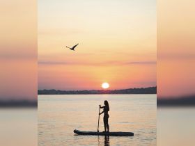A silhouette of a woman paddleboarding at sunset, creating a serene and picturesque scene on the water — perfect for adventure seekers and sunset lovers alike.