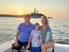 A family enjoys a beautiful sunset on a boat, with a lighthouse in the background — a perfect moment for creating lasting memories on the water.