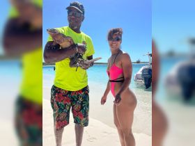 A vibrant scene at the beach featuring a man holding an iguana while a woman poses beside him, showcasing a unique human-animal interaction in a tropical paradise.