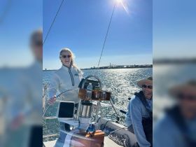 Two individuals enjoy a sunny day sailing, with one at the helm and the other relaxing on the boat — a perfect moment for sailing enthusiasts.