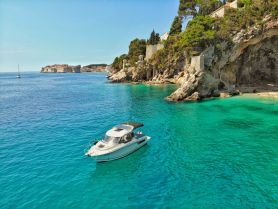 A stunning aerial view of a boat cruising through crystal-clear turquoise waters, with a picturesque coastal backdrop featuring cliffs and historic architecture.