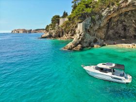 A stunning aerial view of a sleek boat anchored near a rocky coastline, showcasing the vibrant turquoise waters and scenic backdrop of the historic city in the distance.