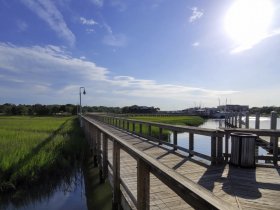 A scenic view of a wooden pier leading into a marsh area, with boats visible in the distance. The image captures the tranquility of the waterfront setting.