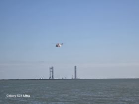An aerial view featuring a helicopter flying over water, with industrial structures in the background. The image lacks clarity and detail.