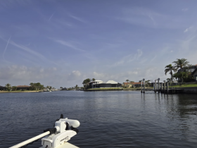 A scenic view of a tranquil waterway with a boat's bow in the foreground, showcasing beautiful waterfront homes and lush palm trees along the banks.