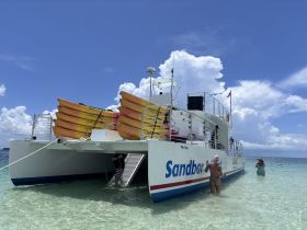 Guests enjoy a sunny day at the sandbar, with a catamaran and kayaks in the background, perfect for water activities and relaxation.