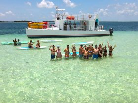 A lively group celebrates at a sandbar, enjoying drinks and paddleboarding in crystal-clear waters — the perfect backdrop for fun-filled aquatic adventures.