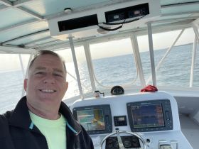 A captain smiles for a selfie in the helm of a boat, showcasing the navigation console and a beautiful view of the water ahead.