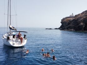 A lively group enjoys a refreshing swim in the clear waters near a sailboat, with a scenic coastline in the background — perfect for summer adventures on the water.