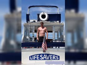 A man stands confidently on the deck of a boat named 'LIFE SAVERS,' showcasing a relaxed day on the water with an American flag waving in the breeze.