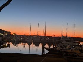 A stunning sunset over a marina, with boats reflecting in the calm waters — a picturesque scene perfect for evening strolls or photography.
