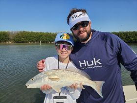 A young angler proudly holds up a redfish alongside an adult, showcasing a successful fishing trip in a scenic waterway.