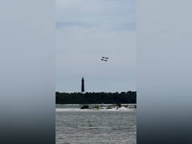 A stunning view of a lighthouse with a formation of jets flying overhead, capturing a perfect moment during an airshow against a scenic backdrop.