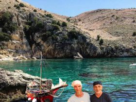 Two men pose in front of a colorful boat anchored near a stunning coastline, showcasing the beauty of the clear waters and rocky landscape — a perfect backdrop for a memorable boating adventure.