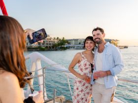 A couple poses for a photo on a boat at sunset, enjoying drinks and capturing memories — perfect for romantic getaways or special celebrations.